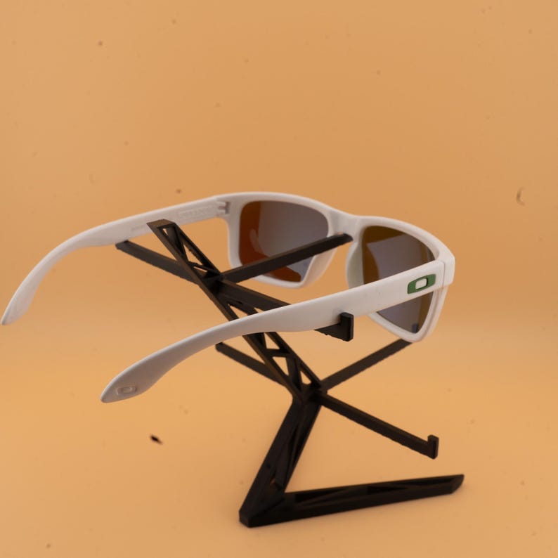 May include: White sunglasses with a rectangular frame and mirrored lenses. The sunglasses are displayed on a black stand against a light orange background. The sunglasses have a green logo on the side.