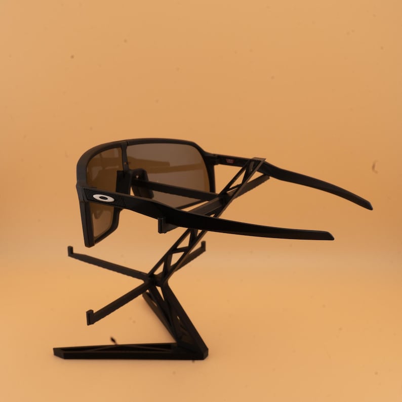 May include: Black sunglasses with brown lenses are displayed on a black stand. The sunglasses have a sporty design with a thick frame and curved temples. The lenses are tinted, and the frame features a small white logo on the side.