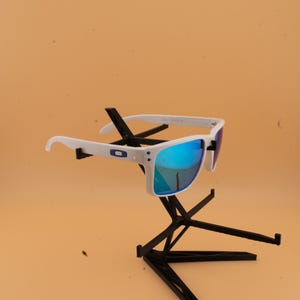 Puede incluir: Gafas de sol blancas con lentes espejadas azules se exhiben en un soporte negro. Las gafas tienen una montura blanca con un logotipo azul en el lateral. Las lentes reflejan un color azul brillante.