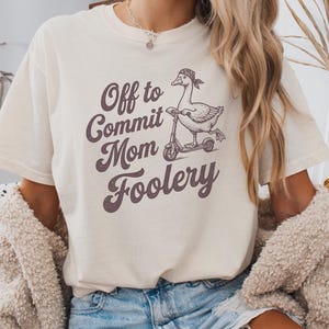 Könnte beinhalten: Ein elfenbeinfarbenes Kurzarm-T-Shirt mit einer dunkelbraunen Grafik einer Gans, die ein Bandana trägt und auf einem Roller fährt. Der in Dunkelbraun gedruckte Text lautet, Off to Commit Mom Foolery. Das T-Shirt hat einen Rundhalsausschnitt.