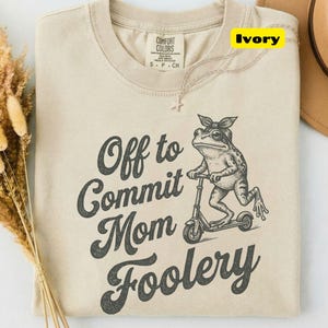 May include: An ivory t-shirt featuring a dark gray graphic of a frog wearing a headscarf and riding a kick scooter. The text reads Off to Commit Mom Foolery in a dark gray font. The shirt has a Comfort Colors brand label at the neck.