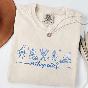 May include: A beige short-sleeve t-shirt featuring a blue graphic print of various orthopaedic medical icons including a knee, a walking boot, crutches, a spine, and a foot, with the word orthopaedics written in a cursive script font below.