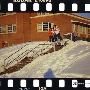 May include: A photograph of a person snowboarding down a snow-covered staircase with metal railings. Two other people are standing nearby. The background features a brick building with windows. The image is framed by a film strip.