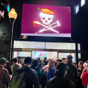 Pu&ograve; includere: Una folla di persone davanti a un edificio di notte. Sopra l'ingresso, un grande schermo mostra un teschio e ossa incrociate con un cappello da pirata e stampelle. Il teschio ha una goccia rossa.