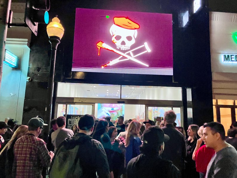Pu&ograve; includere: Una folla di persone davanti a un edificio di notte. Sopra l'ingresso, un grande schermo mostra un teschio e ossa incrociate con un cappello da pirata e stampelle. Il teschio ha una goccia rossa.