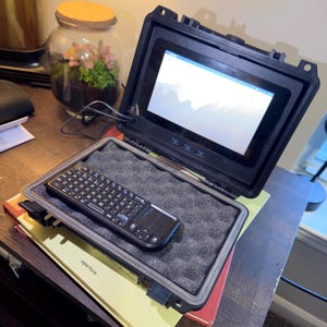 May include: A black portable computer setup with a screen showing a landscape. The screen is in a rugged case, with a keyboard and trackpad on foam inside. A small terrarium is in the background.