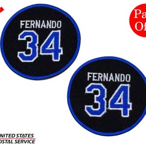 Puede incluir: Dos parches circulares negros con el nombre "FERNANDO" en blanco y el número "34" en azul. Los parches tienen un borde azul. Un círculo rojo indica "Pack Of 2".