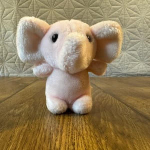 May include: A soft, light pink plush elephant toy with large, floppy ears and dark, round eyes. The stuffed animal features a short trunk and a rounded body, crafted from a fuzzy, textured fabric that gives it a gentle and tactile appearance.