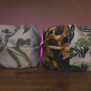 Op de afbeelding: Twee fleece mutsen. De ene muts heeft een witte achtergrond met groene kikker- en bloempatronen. De andere muts heeft een camouflagepatroon met groene en bruine tinten. Beide mutsen zijn aan de onderkant gevouwen.