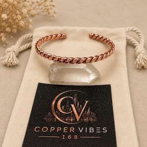 May include: A polished copper cuff bracelet with a twisted rope design resting on a clear quartz crystal. The bracelet is positioned on a beige drawstring pouch featuring a black label with the text COPPER VIBES 168 and a circular logo.