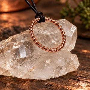 Könnte beinhalten: Ein kupferfarbener Metallring mit gedrehter Seilstruktur, der an einem dünnen schwarzen Lederband hängt. Der kreisförmige Ring ist vor einem klaren, facettierten Quarzkristall positioniert und zeigt den metallischen Glanz sowie das gedrehte Design.