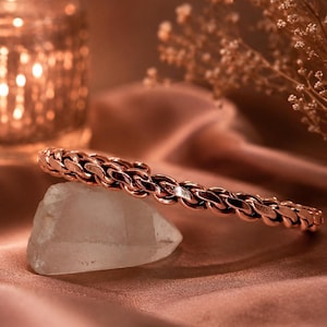 May include: A copper-toned metal bracelet featuring a woven, chain-link design. The bracelet is resting on a translucent, irregular-shaped crystal stone. The metal has a polished, reflective surface with a warm, reddish-brown metallic hue.