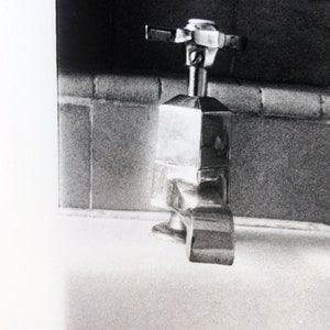 Peut inclure: Un robinet en argent avec une base carr&eacute;e et une poign&eacute;e en forme de croix. Le robinet est mont&eacute; sur un comptoir blanc.