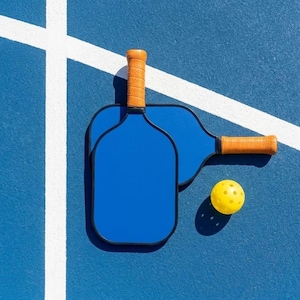 Può includere: Due racchette da pickleball blu con impugnature arancioni e una pallina gialla su un campo blu con linee bianche. Le racchette sono posizionate una accanto all'altra e la pallina è di lato.