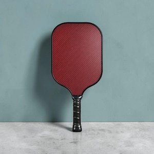 Könnte beinhalten: Ein roter und schwarzer Pickleball-Schläger mit strukturierter Oberfläche. Der Schläger hat einen schwarzen Griff mit Spiralgriff. Der Schläger steht vor einer türkisfarbenen Wand und einem grauen Boden.