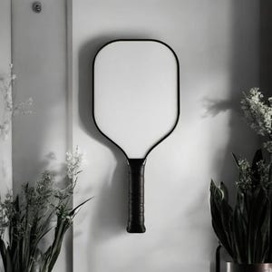 Peut inclure: Une raquette de pickleball noire et blanche avec une face blanche et une poignée noire. La raquette est fixée sur un mur blanc, avec des plantes au premier plan et en arrière-plan.