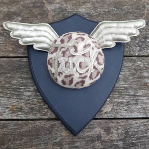 May include: A decorative wall hanging featuring a leopard print fabric sphere with the word "F*ck" embroidered on it. The sphere is attached to a dark blue shield with silver wings.