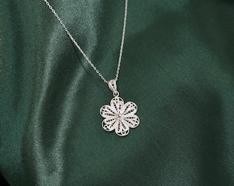 May include: A silver necklace with a delicate flower pendant. The pendant features an openwork design with a small, round center. The necklace chain is thin and hangs against a dark green satin background.