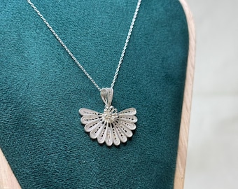 May include: A silver pendant necklace featuring a fan-shaped filigree design. The pendant hangs from a delicate chain and is displayed against a teal background. The intricate openwork design of the pendant is visible.