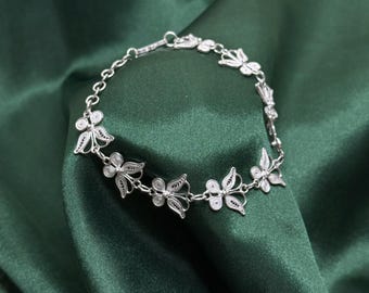 May include: A delicate silver bracelet featuring a series of filigree butterfly charms. The bracelet is displayed on a dark green satin fabric, creating a contrast that highlights the intricate details of the jewelry. The butterfly charms are evenly spaced along the chain.
