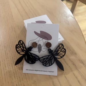May include: Black dragonfly earrings with a cut-out wing design. The earrings have a wooden accent and are displayed on a white card with the text "Add exclusive elegance and beauty to women!"