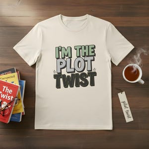 Puede incluir: Camiseta color crema con el texto "I'M THE PLOT TWIST" en verde, gris y negro. La camiseta se muestra sobre una superficie de madera con libros, una taza de té y una etiqueta que dice "plot twist".