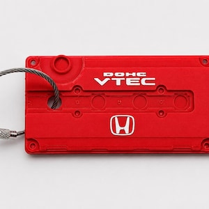 May include: A rectangular red keychain modeled after a Honda engine valve cover. The surface features the white text DOHC VTEC and the Honda logo. A flexible metal cable loop is attached to the left side for securing keys or other items.