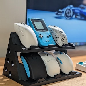 May include: A black two-tiered display stand with hexagonal cutouts on the sides, holding a blue Nintendo Game Boy Color and five computer mice in various colors including white, black, and blue, arranged on the two shelves of the stand.