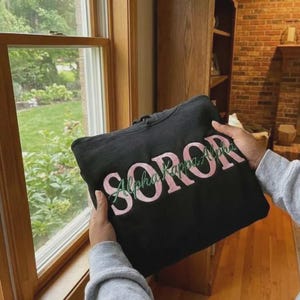 May include: Black hooded sweatshirt with the embroidered text "SOROR Alpha Kappa Alpha" in pink and green. The sweatshirt is held up near a window, showcasing the design.