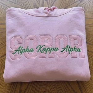 May include: A light pink hooded sweatshirt with the embroidered text "SOROR" above "Alpha Kappa Alpha" in green script. The sweatshirt is folded neatly on a wooden surface.