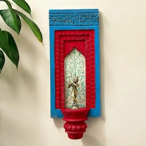 May include: A decorative wall hanging with a blue frame and red interior. The design features a small gold statue on a red pedestal, set against a painted backdrop with a floral pattern. The frame has a carved starburst pattern.