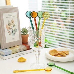 Könnte beinhalten: Eine Sammlung farbenfroher Rührstäbchen mit runden Köpfen in einem klaren, strukturierten Glas. Die Rührstäbchen haben teal, orange, grüne und gelbe Köpfe und Stiele. Eine kleine Sukkulentenpflanze und Bücher befinden sich im Hintergrund.