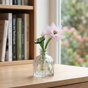 Puede incluir: Un jarrón de cristal transparente con una flor de loto rosa claro y un tallo verde. El jarrón es redondo con un diseño decorativo y se encuentra sobre una superficie de madera. Estanterías en el fondo.