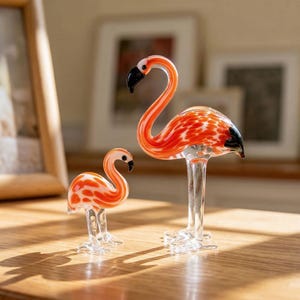 Puede incluir: Dos figuras de flamencos de cristal. El flamenco más grande tiene un cuello largo y curvado y es de color naranja con manchas blancas. El flamenco más pequeño tiene un diseño similar. Ambos tienen picos negros y patas de cristal transparente.