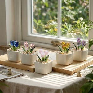 May include: Five small, square, white ceramic pots each hold a glass tulip flower arrangement. The tulips are in shades of blue, pink, yellow, and purple. The pots sit on a wooden board in front of a window.