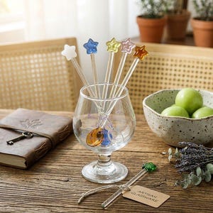 以下が含まれることがあります： さまざまな色の星型のマドラーが入った透明なグラス。マドラーは白、青、黄色、ピンク、オレンジです。グラスは、ノート、青リンゴのボウル、ラベンダーの小枝の隣の木製のテーブルに置かれています。「ARTISAN GLASSWARE」と書かれたタグが付いています。