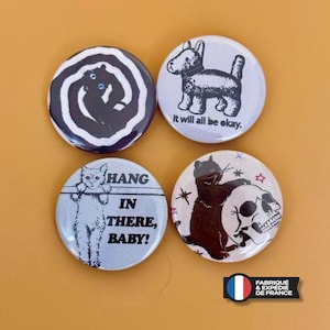 Può includere: Quattro spille rotonde con illustrazioni grafiche. Una mostra un gatto nero in una spirale bianca, un'altra un cane con il testo 'it will all be okay.', una terza un gatto con 'HANG IN THERE, BABY!', e l'ultima un gatto con un teschio.