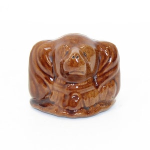 Puede incluir: Una pequeña figurita de cerámica marrón de un perro con la cabeza gacha y las orejas dobladas sobre los ojos. El perro lleva un collar marrón y tiene una nariz marrón.