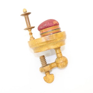 Puede incluir: Un cojín de alfileres de madera con una parte superior de tela roja y una abrazadera para sujetarlo a una mesa o superficie.