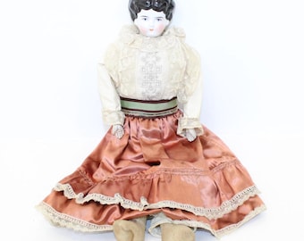 Può includere: Bambola antica in porcellana con capelli neri, vestita con una blusa di pizzo bianco con un nastro verde e una gonna di raso color pesca con bordatura di pizzo.