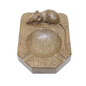 May include: A rectangular wooden ashtray featuring a carved mouse figure on the top surface. The piece has a central circular basin and a long, carved tail extending down the side. The wood displays a visible grain pattern throughout the object.