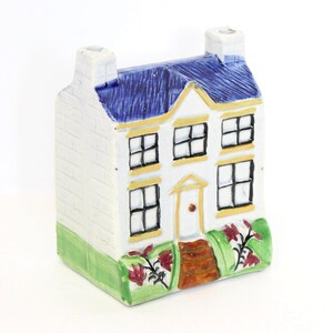 Puede incluir: Una pequeña figura de casa de cerámica pintada a mano. La casa tiene un exterior blanco con un tejado azul, marcos de ventanas amarillos y una base verde con flores rojas. Tiene dos chimeneas y una puerta principal.