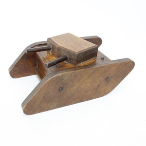May include: A wooden toy tank with a brown finish. The tank has a small turret with two cannons on top. The tank is on a white background.