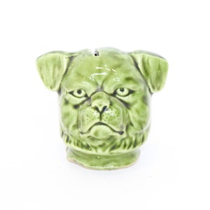 May include: Green ceramic pug dog head figurine. The pug has a serious expression with a furrowed brow and a slightly open mouth.