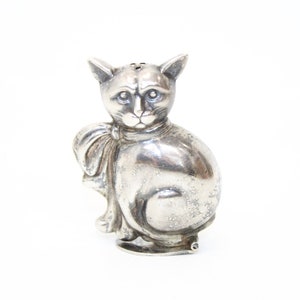 Könnte beinhalten: Eine silberne Katzenfigur mit einer Schleife um den Hals. Die Katze sitzt mit leicht geneigtem Kopf. Die Figur ist ein Salz- und Pfefferstreuer.