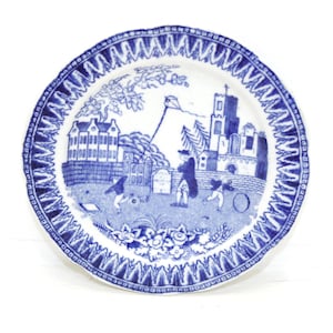 May include: A blue and white decorative plate with a detailed landscape scene. The plate has a blue zigzag border. The central image shows figures flying a kite near buildings and trees, reminiscent of a historical print.