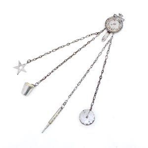 Op de afbeelding: Een zilverkleurige chatelaine-broche met een rond bovendeel voorzien van een kroonmotief. Aan de basis hangen vier fijne metalen kettinkjes, elk bevestigd aan een andere bedel: een vijfpuntige ster, een vingerhoed, een naaldenkoker en een ronde schijf.