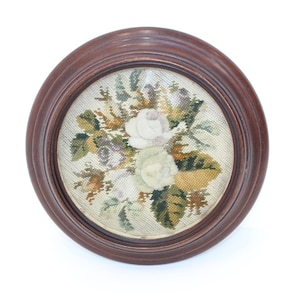 May include: A framed needlepoint embroidery featuring a bouquet of white roses with green and brown leaves on a white background.