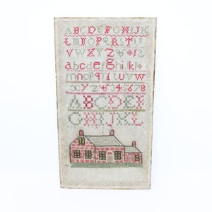 May include: A framed sampler with a house design and the alphabet in red and green thread. The sampler is on a white background.