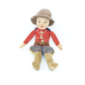 May include: A vintage plush doll dressed in a red uniform with a brown belt and a brown hat. The doll is sitting with its legs crossed and has a painted face.